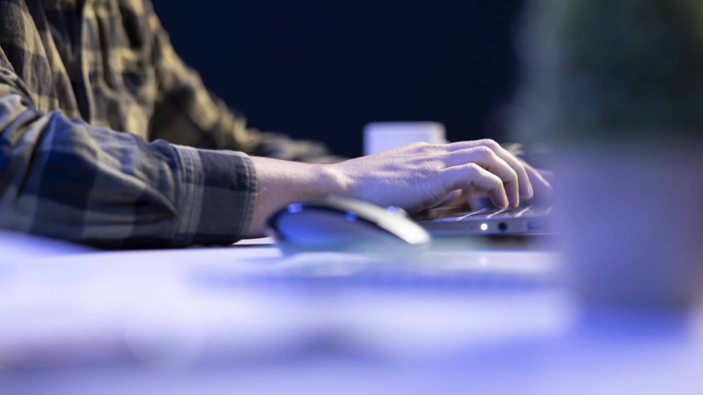 Closeup shot of male individual using laptop keyboard and mouse at office desk to surf the internet. Person typing on digital device, finishing tedious paperwork.