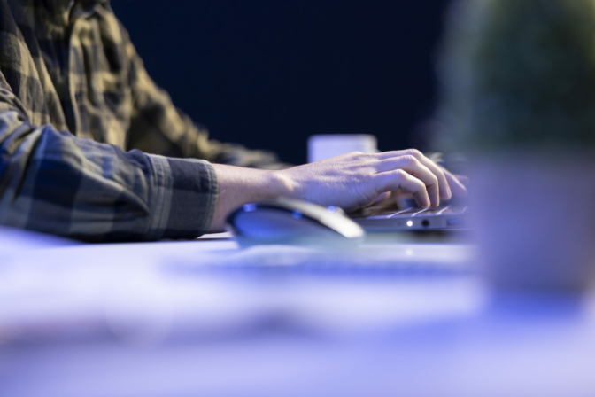 Closeup shot of male individual using laptop keyboard and mouse at office desk to surf the internet. Person typing on digital device, finishing tedious paperwork.